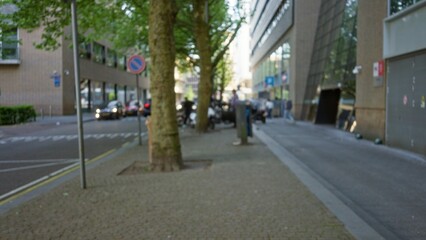 Blurred view of an urban european city street with defocused background, showcasing cars, trees, and pedestrians in a town setting with a bokeh effect.