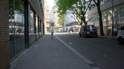 Urban street scene with blurred figures in a european city; woman walking in defocused background amidst modern architecture under blue sky and green trees.
