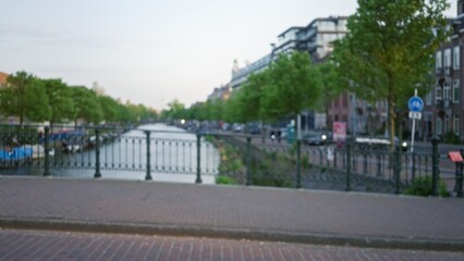 Blurred view of amsterdam canal with defocused trees, urban street, and classic buildings, capturing a tranquil evening in the netherlands.