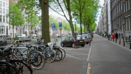 Blurred bicycles line a serene canal in amsterdam, netherlands, with cars parked under lush green trees, creating a classic european street scene with defocused elegance and charm.