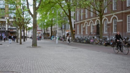 Fototapeta premium Blurred view of european city street with people walking and cycling on cobblestone path alongside historic buildings and green trees, creating a bokeh effect.
