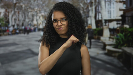Woman with curly hair and glasses stands outdoors on a city street making hand gestures while wearing a black top. the urban setting is lively and bustling with activity.