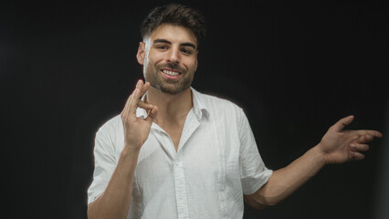Man gesturing ok sign with right hand and pointing finger with left hand in studio against black backdrop; confidence approval.