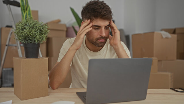 Man gestures with raised hands over laptop on wooden table and new moving boxes inside building; confusion.