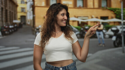 Young hispanic woman pointing finger and smiling in a white crop top and denim jeans on a city street lined with scooters and cafe terrace; joy curiosity adventure.