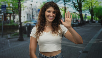 Woman waves hand on amsterdam street by canal and trees, smiling in white crop top and jeans while facing camera; friendly greeting joy.