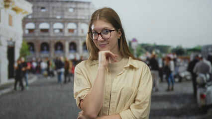 Fototapeta premium Young blonde woman wearing glasses in beige shirt touches chin with hand in street by coliseum; curiosity.