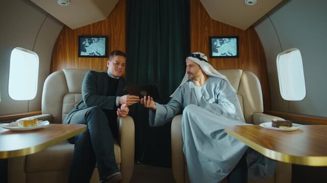 Diverse business partners having a conversation on a luxurious private jet, with an arab businessman in traditional clothing using a tablet to discuss a contract during an international flight