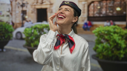 Blonde air hostess wearing cap and scarf covers eye with hand while laughing on bustling city street; joy.