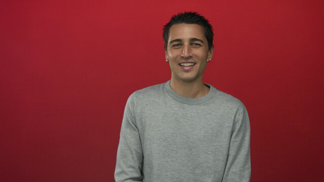 Young hispanic man smiling confidently against a vibrant red background wearing a casual grey sweater, conveying a relaxed and friendly demeanor.