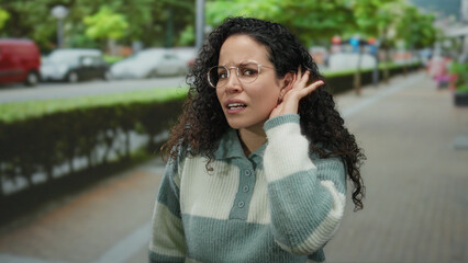 Fototapeta premium Hispanic woman with curly hair and glasses stands outdoors on a street, gesturing to listen with her hand at her ear, surrounded by greenery and vehicles.