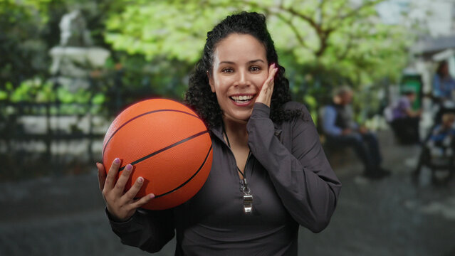 Woman holding basketball in park setting with bright greenery, expressing enthusiasm and joy, suggesting outdoor sports coaching or recreation activities.