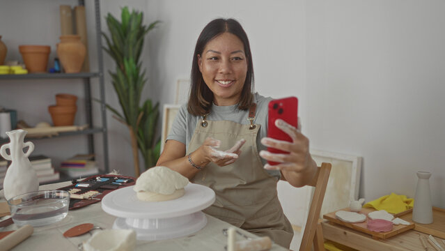 Woman holding red smartphone and waving clay covered hand at camera in pottery studio with wheel and tools on table; joy creative connection.