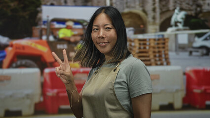 Woman in apron holds up two fingers peace sign in front of construction barriers and forklift at building; confidence peace.