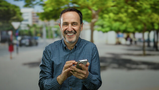 Man smiling with smartphone in urban park setting with green trees and buildings blurred in background.