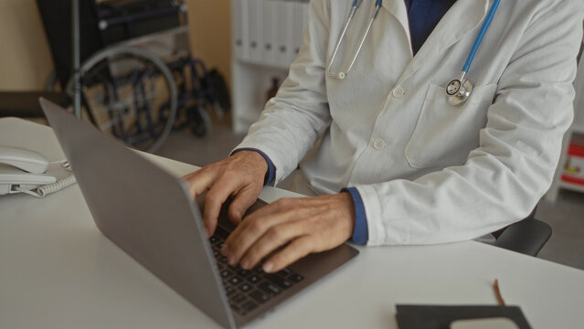 Doctor working in hospital office typing on laptop with stethoscope around neck focused on patient care surrounded by medical equipment and supplies.
