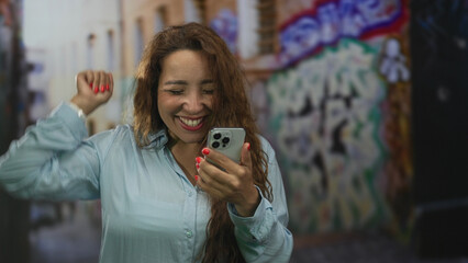 Woman holding smartphone, smiling and waving hand on a street with graffiti while checking messages...
