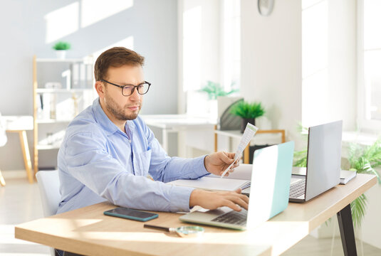 Serious young man in glasses working at desk with two laptops and documents in bright office space. Focused multitasking company employee analyzing papers and typing, managing tasks or reviewing data.