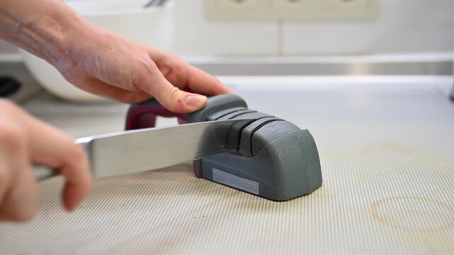 Close-up of female hand sharpening metal kitchen knife using manual sharpener on countertop, home kitchen interior, safe cooking process.