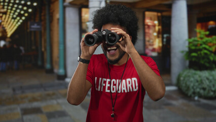 African american man in lifeguard shirt holding binoculars with a bright smile standing on a lively...