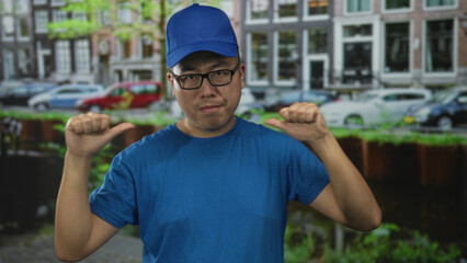 Man in blue cap and t shirt thumbs pointing to chest on a street with parked cars and canal; self belief confidence.