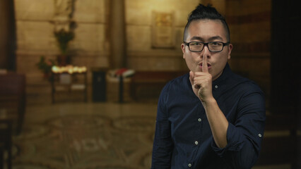 Man finger to lips for silence in church interior wearing glasses and navy shirt; reverence...