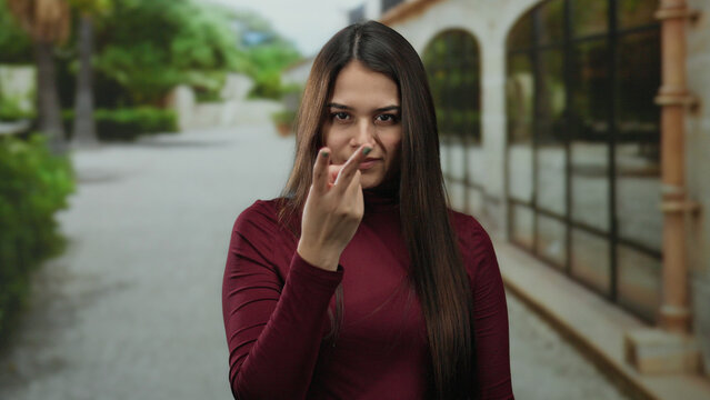 Hispanic woman making watchful and threat gestures on a street, wearing a burgundy shirt, conveying a serious expression in an outdoor setting with greenery.