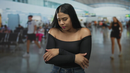Young latina woman in black off shoulder top hugs arms across chest at busy airport terminal; unease.