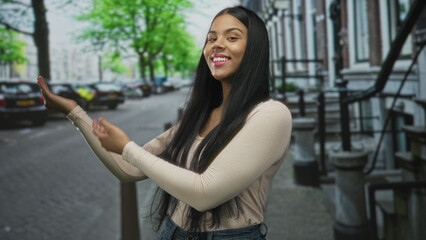 Latin young woman gestures arms with open palms amid sunny urban tree lined street by parked cars; warmth invitation friendliness openness.