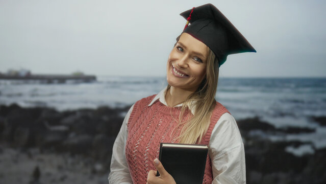 Young woman celebrating graduation at seaside holding book with blonde hair and cap smiling outdoors at the beach with ocean waves in the background