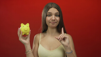 Girl holds yellow piggy bank in left hand and points finger to bank in red studio scene; skepticism.