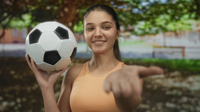 Woman caucasian teenager extends hand to camera holding soccer ball in park under green trees; youthful sport confidence.