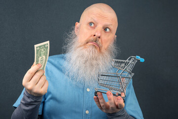 Man with a beard holding dollar bill and shopping cart contemplating spending choices