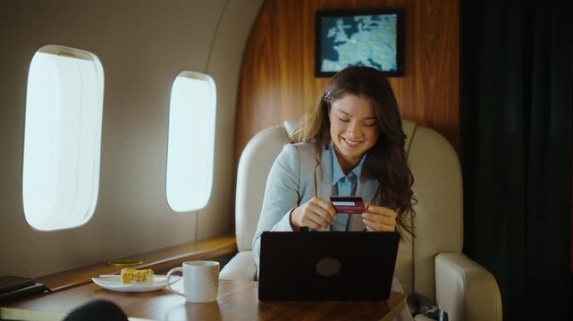Happy successful businesswoman booking tickets or shopping online with a credit card and laptop while flying first class, enjoying her luxurious trip and celebrating her purchase with a smile