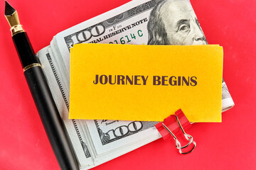 A stack of United States currency, a yellow notepad with the words Journey Begins, a red binder clip, and a black fountain pen lay on a red surface
