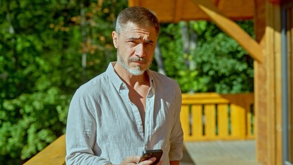 Older man standing on balcony at home using cell phone in warm morning sunlight. Portrait of middle aged casual male looking at camera using social media on mobile.