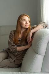 Obraz premium Cute young woman sitting on sofa at home, looking out window. Relaxed lifestyle, indoor leisure, cozy home, thoughtful, casual everyday moment.