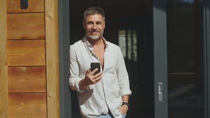 Mid adult man standing at balcony door drinking morning coffee on terrace at home. Vacation in mountain cabin house. Freedom, lifestyle and getaway .