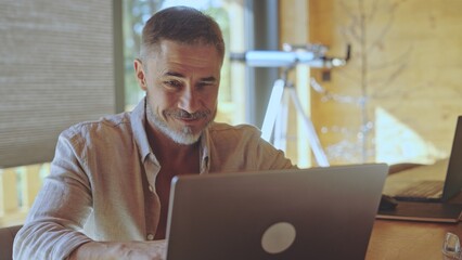 Middle aged man sitting at desk remote working with laptop in home office. Businessman on vacation managing business online. Happy casual mid adult male smiling. Digital technology.