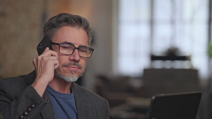 Serious businessman working in home office talking on phone call. Middle aged, older male in glasses concentrating, using laptop and mobile. Telecommunication, entrepreneurship.