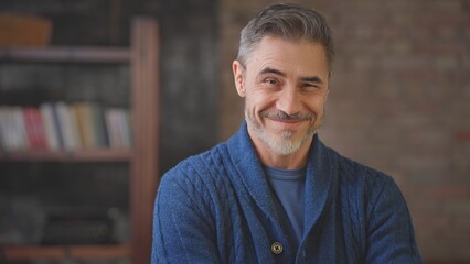 Portrait of happy casual middle aged man at home in loft room. Senior male with grey hair looking at camera wearing sweater, smiling. Books and typewriter in background.
