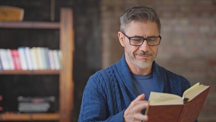 Happy casual middle aged man reading book at home in loft room. Senior male with grey hair in eyeglasses, smiling. Books and typewriter in background, lifestyle of smart older guy.