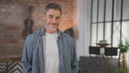 Portrait of happy casual middle aged man standing at home in loft living room. Senior male with grey hair in denim shirt looking at camera, smiling.