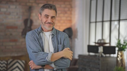 Portrait of happy casual middle aged man standing at home arms crossed in loft living room. Senior male with grey hair in denim shirt looking at camera, smiling.