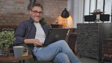 Senior businessman sitting in armchair in living room remote working in home office with laptop. Portrait of happy, confident middle aged man, smiling. Entrepreneur managing business.