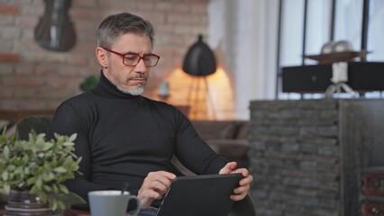 Senior businessman sitting in armchair in living room remote working in home office with digital tablet. Middle aged man in turtleneck and glasses. Entrepreneur managing business.
