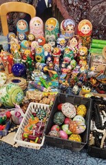 A vibrant market featuring a variety of painted matryoshka dolls, wooden toys, and decorated eggs displayed on a fabric-covered table. An atmosphere of traditional craftsmanship and folk art.