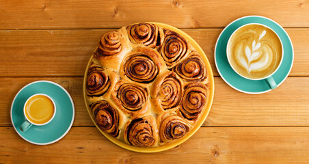 Homemade cinnamon rolls from yeast dough and two cups of coffee cappuccino and espresso on wooden table. Top view.