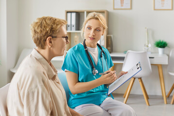 Obraz premium Discussion of nurse and senior woman on doctors appointment in clinic. Professional medical worker in uniform talking, looking to elderly patient on consultation, holding clipboard with notes