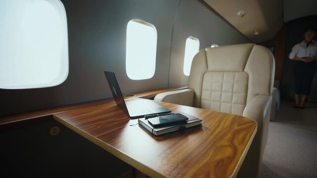 Elegant and modern interior of a private business jet with a comfortable leather seat, a wooden table with a laptop, and a flight attendant standing in the background during the flight